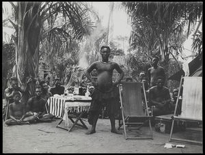 Kot aPe, the Nyimi (chief) of Mushenge (Nsheng) standing next to European style table and deck-chairs, crowd seated on ground behind him Kot aPe, the Nyimi (chief) of Mushenge (Nsheng) standing next to European style table and deck-chairs, crowd seated on ground behind him