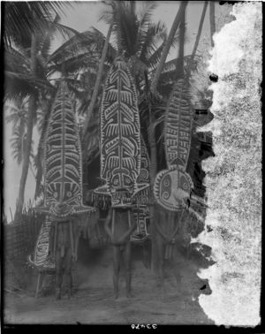 Three men wearing large painted masks, other masks behind them leaning against outside of hut (see also RAI No. 33473, 33499) Three men wearing large painted masks, other masks behind them leaning against outside of hut (see also RAI No. 33473, 33499)