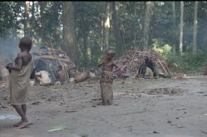 A child (Tutu) standing in the centre of a clearing playing an upright carved wooden drum. A child (Tutu) standing in the centre of a clearing playing an upright carved wooden drum.